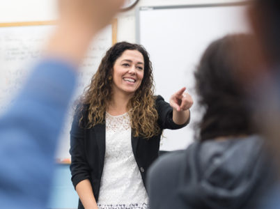Teacher stood at the front of class, selecting a student to answer a question. A student with their hand up is in the foreground.