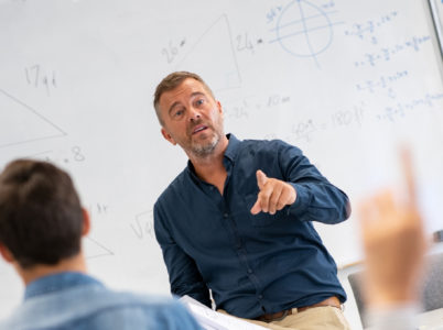 Teacher at the front of class pointing at a student in the foreground with her hand up