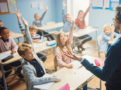 Photo of a teacher at the front of a classroom asking a question; many students have their hands up in the background
