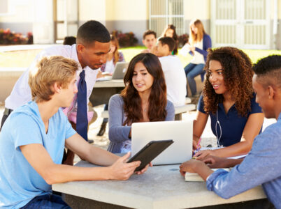 Students Working On Campus With Teacher Using Electronics